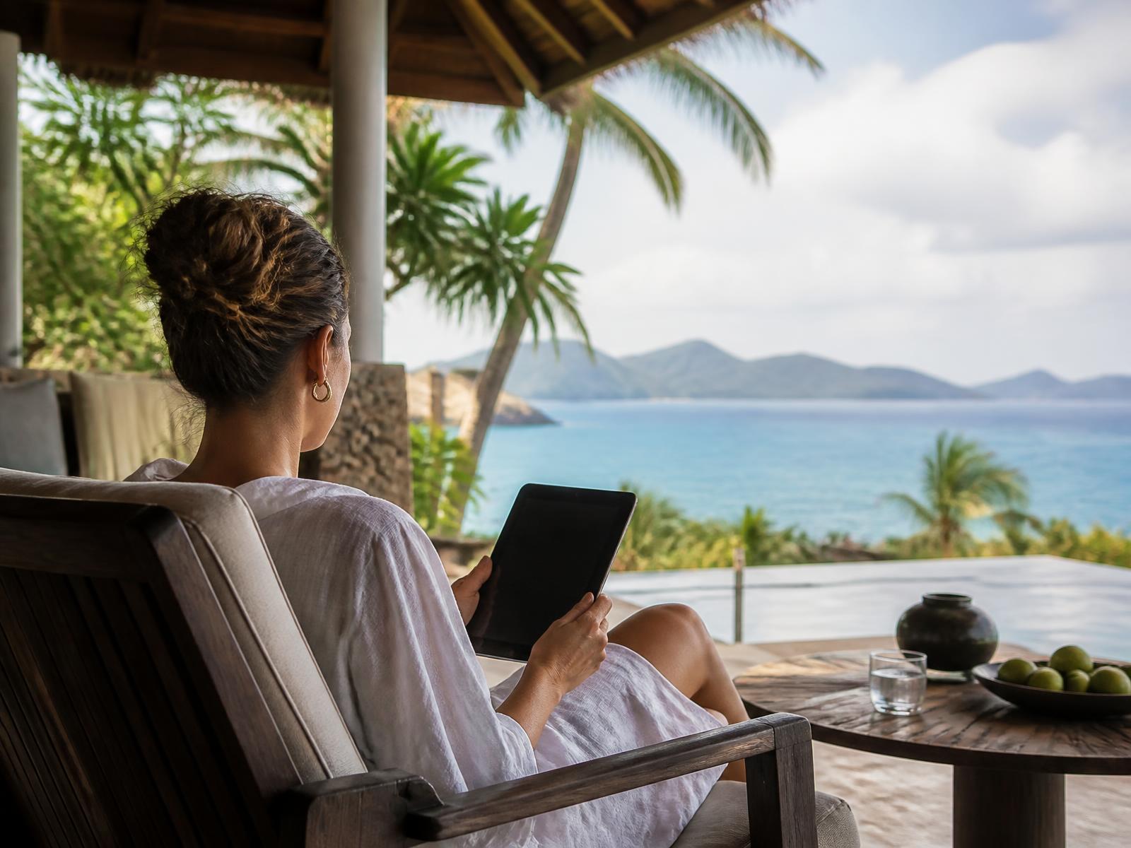 Guest on a shaded resort terrace browsing on a tablet with an ocean view